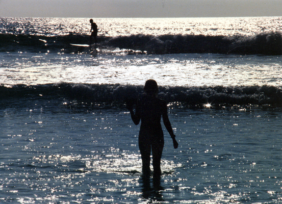 A young woman stands in the shallows of the ocean watching a surfer ride a wave. 