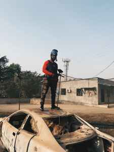 A man holding a paintball gun stands on a burnt out car. 